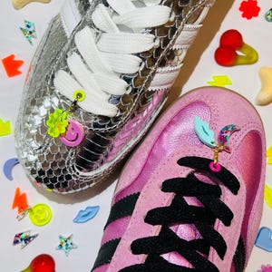 May include: A silver metallic snakeskin-textured sneaker with white laces and a neon yellow flower and pink smiley face charm, alongside a pink metallic sneaker with black laces and a light blue lip and glittery crescent moon charm.