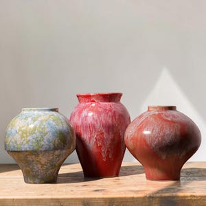 May include: Three ceramic vases with distinct glazes. The left vase has a blue and green mottled pattern. The centre vase is a tall, rounded red vessel with white drip glaze. The right vase is a wide, spherical terracotta-toned piece with white glaze.