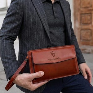 Op de afbeelding: Een rechthoekige bruine leren clutch met een polsband en een voorflap met het in reliëf aangebrachte logo en de tekst velarocrafts. De tas heeft zichtbare stiksels langs de randen en een gladde afwerking, horizontaal vastgehouden.