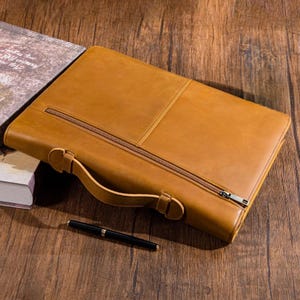 May include: A tan leather briefcase with a zipper closure and a handle. A black pen rests on the wooden surface next to the briefcase. The briefcase has a rectangular shape and a smooth texture.