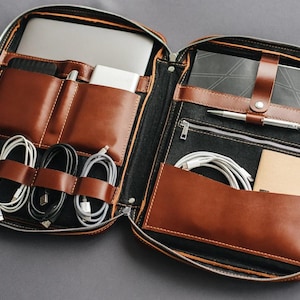 May include: A brown leather zipped organiser case featuring multiple interior compartments. The left side holds a laptop, a power bank, and four coiled charging cables. The right side contains a notebook with a strap, a pen, and a pocket with a cable.