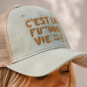 Op de afbeelding: Lichtblauwe truckerpet met een mesh achterkant en een gebogen klep. De pet heeft de geborduurde tekst "C'EST LA F***ING VIE!" in bruine letters. De klep heeft kleine, gelijkmatig verdeelde gaatjes.