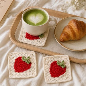 May include: Three square crochet coasters featuring a cream-colored background with a central red strawberry motif and green leaves. One coaster sits under a white ceramic mug filled with green matcha latte, while two others are placed nearby.