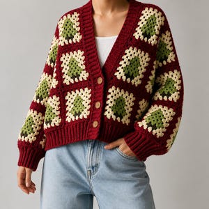 Könnte beinhalten: Eine langärmelige Häkelstrickjacke mit Granny-Square-Muster in Dunkelrot, Creme und Grün. Das Kleidungsstück hat einen V-Ausschnitt, einen gerippten dunkelroten Saum und drei runde Holzknöpfe auf der Vorderseite, kombiniert mit einer hellblauen Jeans.