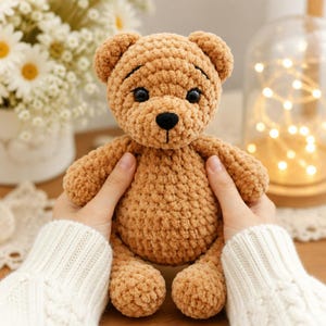 May include: A light brown crochet teddy bear made of soft, chunky yarn. The bear features a round head with small ears, two black plastic eyes, and a black embroidered nose and mouth. The body is plush and textured with a consistent stitch pattern.
