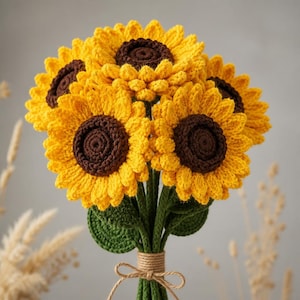 Könnte beinhalten: Ein Strauß aus fünf gehäkelten Sonnenblumen mit leuchtend gelben Blütenblättern und dunkelbraunen Mitten. Die Blumen haben grüne gehäkelte Stiele und Blätter, und die Stiele sind mit einer hellbraunen Schnur zu einer einfachen Schleife gebunden.