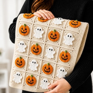 May include: A cream-colored crochet blanket featuring a grid of square panels. Each square is decorated with an appliqued motif of either an orange jack-o'-lantern with a green stem or a small white ghost with black eyes, arranged in an alternating pattern.