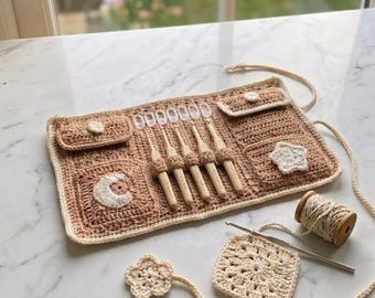 May include: A beige crochet organizer roll featuring five wooden crochet hooks, two small pockets with button closures, and decorative white moon and star appliques. Includes a matching square granny square, a flower motif, and a spool of yarn.