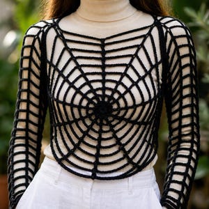 Puede incluir: Un top de ganchillo negro con un patr&oacute;n de telara&ntilde;a sobre una camisa de cuello alto color crema de manga larga y pantalones blancos. La capa negra tiene un dise&ntilde;o de red circular en el pecho y un patr&oacute;n de rejilla en las mangas largas.