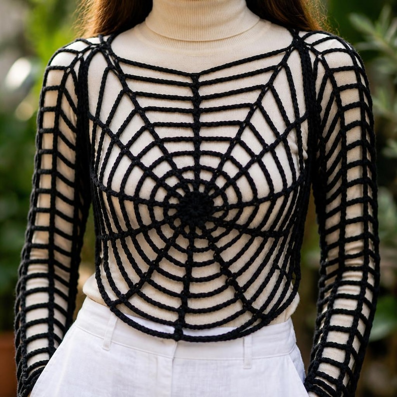Puede incluir: Un top de ganchillo negro con un patr&oacute;n de telara&ntilde;a sobre una camisa de cuello alto color crema de manga larga y pantalones blancos. La capa negra tiene un dise&ntilde;o de red circular en el pecho y un patr&oacute;n de rejilla en las mangas largas.