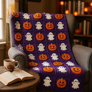 May include: A purple crochet blanket featuring a repeating pattern of orange jack-o'-lanterns and white ghosts in individual square blocks. The blanket is draped over a chair, with a ceramic mug featuring a pumpkin illustration and an open book nearby.