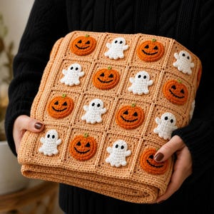 May include: A square crochet blanket featuring a grid of light tan squares, each decorated with either a bright orange jack-o'-lantern with a black stitched face or a small white ghost with black eyes, creating a repeating Halloween-themed pattern.