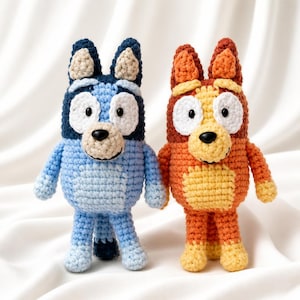 May include: Two handmade crochet plush toys representing cartoon dogs. The left figure is dark and light blue with white eye patches. The right figure is orange and yellow with white eye patches. Both have black noses, small black eyes, and tall ears.