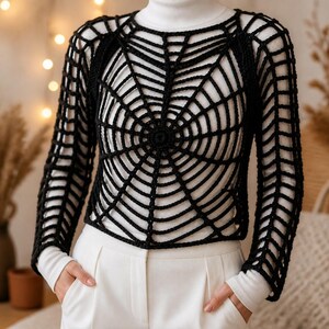 Puede incluir: Una blusa de crochet negra de manga larga con un patr&oacute;n de telara&ntilde;a prominente en la parte delantera y las mangas. La prenda se lleva sobre una camiseta blanca de cuello alto de manga larga y pantalones blancos, creando un estilo superpuesto.