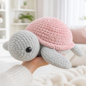 May include: A plush crochet turtle toy featuring a soft, textured light gray head and flippers, paired with a rounded, dusty pink shell. The turtle has a single visible black plastic eye on the side of its head, crafted from thick yarn.