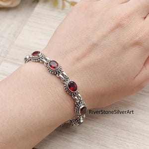 Op de afbeelding: Een zilveren schakelarmband met meerdere ovaalvormige, dieprode granaatedelstenen in bewerkte metalen zettingen. De armband heeft een herhalend patroon van gepolijste zilveren schakels die elke steenzetting met elkaar verbinden.