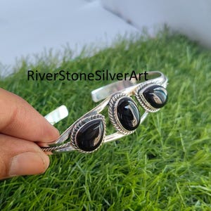 Può includere: Un bracciale a polsino in argento con tre pietre di onice nera. Le pietre sono a forma di goccia e ovali, incastonate in una cornice d'argento decorativa. Il bracciale è esposto su erba verde. Il testo "RiverStoneSilverArt" è visibile.