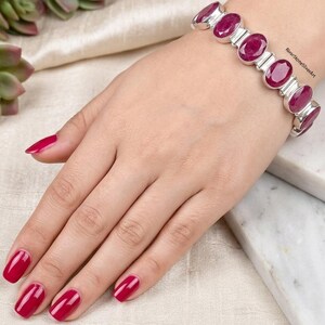 Peut inclure: Un bracelet en argent composé d'une série de pierres précieuses rubis rose foncé de forme ovale, serties dans des maillons en métal poli. Le bracelet est porté au poignet et le texte RiverStoneSilverArt est visible en petites lettres noires.