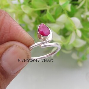 Può includere: Un anello in argento con una pietra preziosa rosa intenso a forma di goccia incastonata in una fascia di metallo lucido. L'anello presenta un design aperto in stile bypass in cui le estremità della fascia si sovrappongono. Il testo RiverStoneSilverArt è visibile.