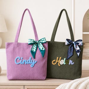 May include: A purple corduroy tote bag with the name Cindy embroidered in blue and a green checkered ribbon bow, and an olive green corduroy tote bag with the name Helen embroidered in yellow and blue and a patterned ribbon bow.
