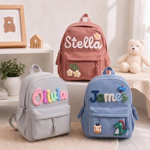 May include: Three corduroy backpacks in dusty rose, light grey, and light blue. The rose bag features the name Stella and a flower patch. The grey bag displays the name Olivia in multicoloured letters. The blue bag shows the name James with dinosaur and giraffe patches.
