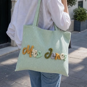 May include: A light green corduroy tote bag featuring the text Mrs Jones in raised, multi-colored knitted yarn letters. The bag has two shoulder straps and a textured ribbed fabric surface, displaying the name in shades of yellow, cream, and green.