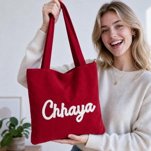 May include: A deep red corduroy tote bag featuring two long shoulder straps and the name Chhaya embroidered in white cursive script across the centre of the front panel. The bag has a rectangular shape and a textured fabric surface.