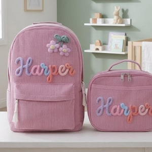 Könnte beinhalten: Ein Rucksack und eine passende Lunchtasche aus rosa Kordstoff. Beide Artikel tragen den Namen Harper in erhabenen, mehrfarbigen Garnbuchstaben. Der Rucksack ist mit einer dekorativen Garnblume und weißen Quasten an den Reißverschlüssen versehen.