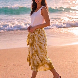 May include: A white sleeveless tank top paired with a mustard yellow and white tie-dye sarong skirt. The skirt features a knotted side detail and falls to mid-calf length. The ensemble is completed with simple brown flip-flop sandals.