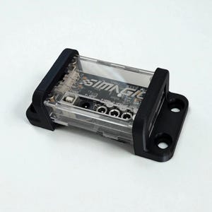 Puede incluir: Un módulo de control electrónico rectangular con una carcasa de plástico transparente y soportes de montaje negros a cada lado. La placa de circuito interna es visible, con el texto SIMAGIC impreso en blanco y varios puertos y conectores.