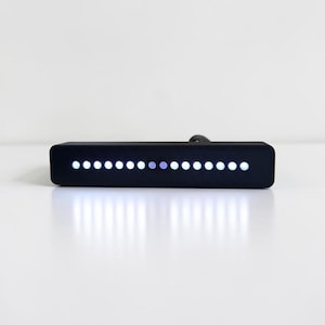 May include: A rectangular black electronic device featuring a horizontal row of sixteen small circular LED lights. The lights emit a bright white glow, with one light appearing slightly dimmer or purple-tinted. A black power cord extends from the back.