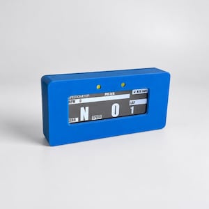 May include: A rectangular blue digital racing dashboard display featuring a screen with white text showing SPEEDOMETER, RPM 0, POS N/A, N/A Laps, GEAR N, SPEED 0, and LAP 1. Two small yellow circular indicator lights are positioned above the screen.