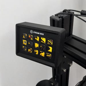 May include: A black Stream Deck control panel with 15 illuminated buttons. The buttons display various icons and patterns in yellow and orange. The Stream Deck is mounted on a black metal frame.