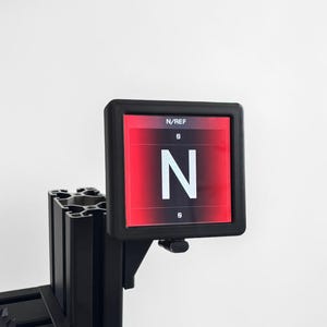 Könnte beinhalten: Ein schwarzer elektronischer Bildschirm mit dem Text "N/REF" und dem Buchstaben "N" in Weiß auf rotem Hintergrund. Der Bildschirm ist auf einem schwarzen Metallrahmen montiert.