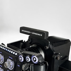 May include: A black racing steering wheel controller featuring a horizontal LED rev light strip, multiple illuminated circular buttons labeled with icons, and metallic rotary dials. The device includes text labels such as Traction, Page, and Adjust.