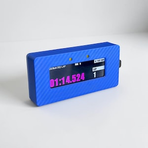 Può includere: Un cronometro digitale rettangolare blu con texture in fibra di carbonio. Lo schermo mostra il testo ESTIMATED LAP, POS 0, 0.0 Laps e LAP 1, con il tempo 01:14.524 visualizzato in grandi cifre rosa. Sono visibili due piccole luci circolari gialle.