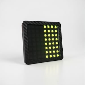 May include: A black square device with a carbon fiber texture frame. The device has a grid of yellow LED lights on the right side, and a dark green grid on the left side. The device is angled slightly.
