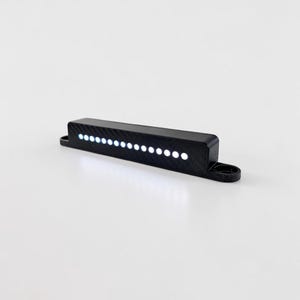 Puede incluir: Un componente electrónico negro y rectangular con una fila de catorce pequeñas luces LED circulares blancas integradas en su parte frontal. La carcasa tiene un acabado texturizado tipo fibra de carbono y pestañas de montaje en los extremos.