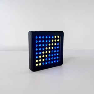 May include: A square black electronic display device featuring an eight-by-eight grid of small circular LED lights. The lights are illuminated in a combination of blue and yellow, with the yellow lights forming a diagonal arrow pattern.