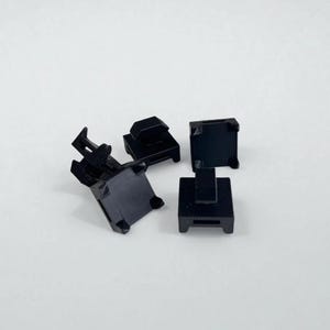May include: A collection of four small, black plastic mounting clips. Each clip features a square base with a central protruding tab and a slotted opening on the side, designed for securing cables or wires in a fixed position on a flat surface.