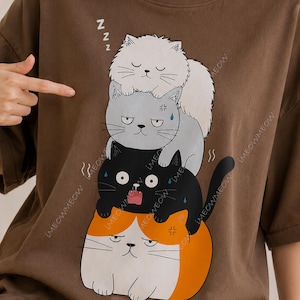 May include: Brown t-shirt featuring a stack of cartoon cats with various expressions. The top cat is white and sleeping, the middle cat is gray and angry, the bottom cat is black and surprised, and the base cat is orange and white and angry. The text "MEOWMEOW" is printed on the shirt.