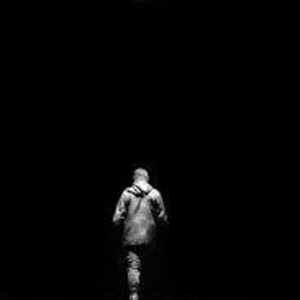 May include: A grayscale image of a person walking away from the camera, under a single overhead light. The person is wearing a hooded jacket and pants, with the background completely dark.