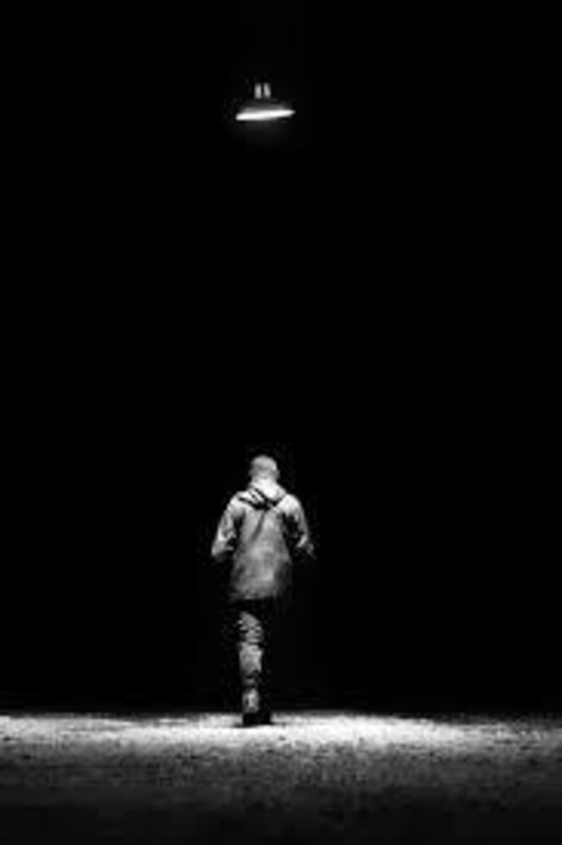 May include: A grayscale image of a person walking away from the camera, under a single overhead light. The person is wearing a hooded jacket and pants, with the background completely dark.