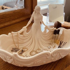 May include: A cream-coloured decorative jewellery dish featuring a sculpted woman in a flowing gown. The dish holds various rings, earrings, a necklace, and small perfume bottles. The dish is placed on a wooden surface.