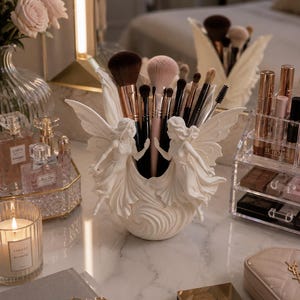 May include: A white, fairy-themed makeup brush holder filled with various brushes. The holder features two sculpted fairies with outstretched wings. Other items on the vanity include perfume bottles, a candle, and a Dior book.