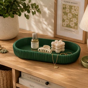 Puede incluir: Una bandeja ovalada verde oscuro y acanalada contiene un frasco de perfume, una pinza para el pelo blanca, una vela de burbujas y joyas de oro. Un pequeño plato verde y una impresión enmarcada también están sobre la superficie de madera. Una lámpara y una planta en maceta están en el fondo.