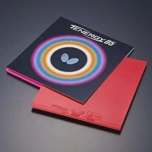Puede incluir: Una goma de tenis de mesa roja con una funda de embalaje negra que muestra el texto TENERGY 05, un logotipo de mariposa central y un vibrante patr&oacute;n circular de degradado de arco&iacute;ris sobre un fondo oscuro.