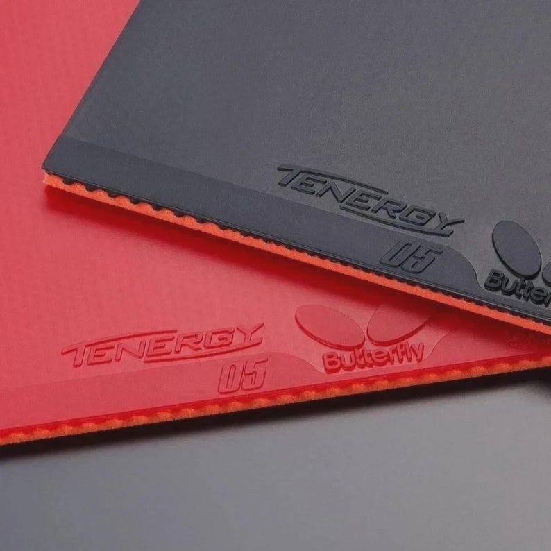 Puede incluir: Dos gomas de tenis de mesa, una negra y otra roja, apiladas una sobre otra. Cada hoja presenta el texto en relieve TENERGY 05 y el logotipo de la marca Butterfly. Los bordes muestran una capa de esponja naranja texturizada bajo la superficie lisa.