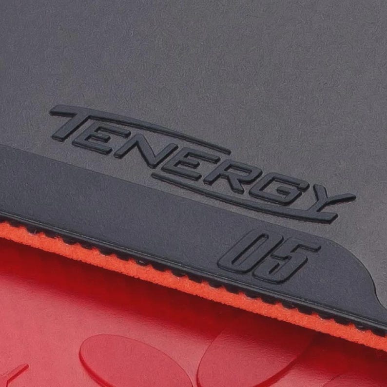 Puede incluir: Una goma de tenis de mesa negra con el texto en relieve TENERGY 05. La goma presenta una superficie superior negra mate con un borde texturizado, sobre una base de esponja naranja brillante, situada encima de una superficie de goma roja lisa.