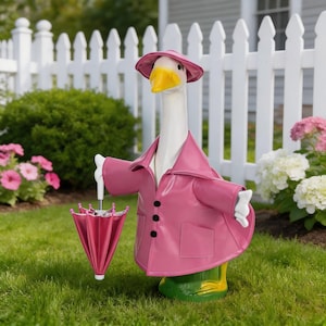 May include: A whimsical garden goose statue wearing a pink raincoat and hat, holding a matching pink umbrella. The goose has a yellow beak and green feet, standing on a grassy lawn in front of a white picket fence and flowers.